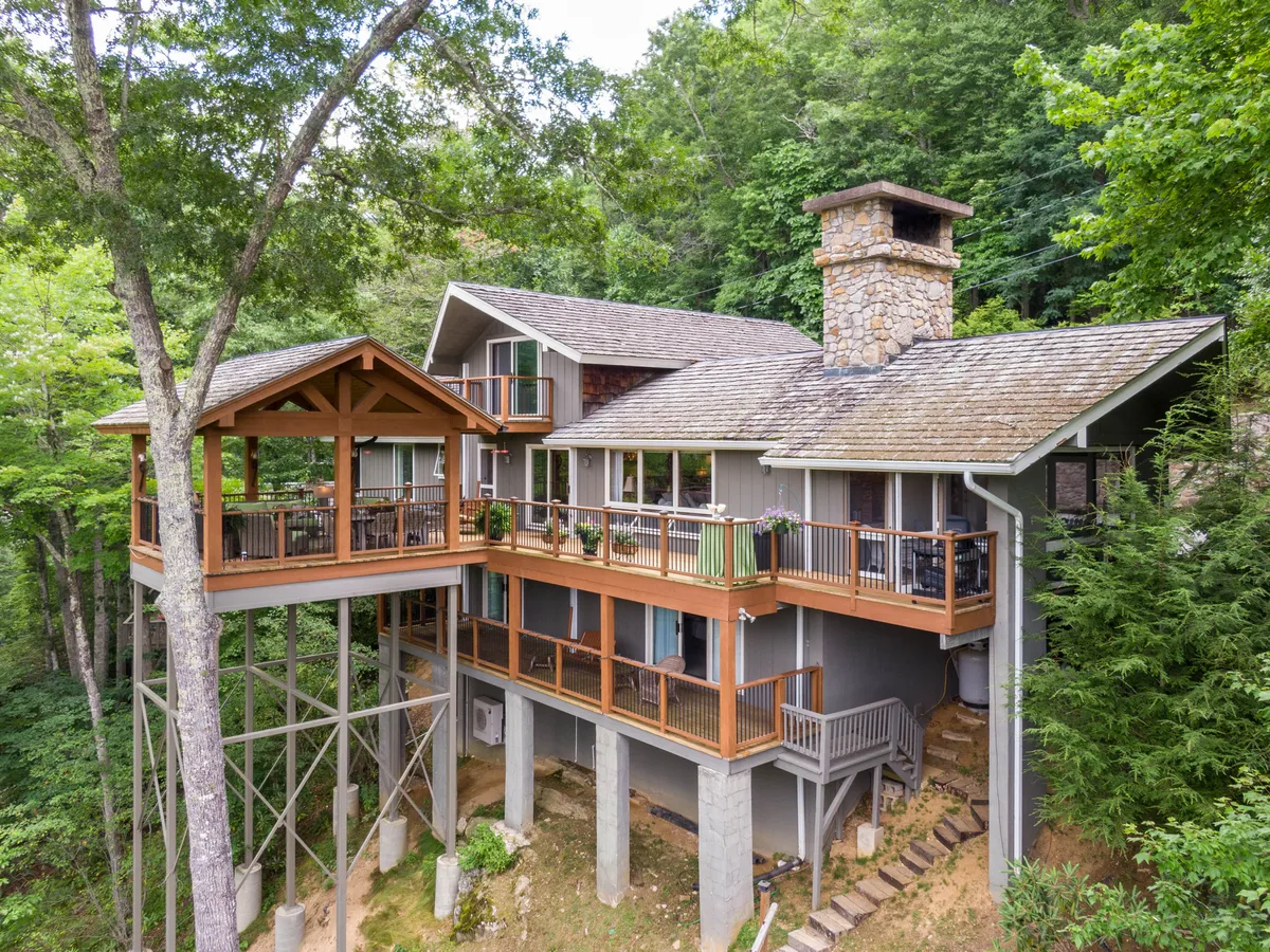 A view of the home's stone chimney, elevated deck, expansive windows, and surrounding trees, showcasing a peaceful and cozy exterior. The scene suggests opportunities for outdoor dining, group relaxation, and sunset watching.