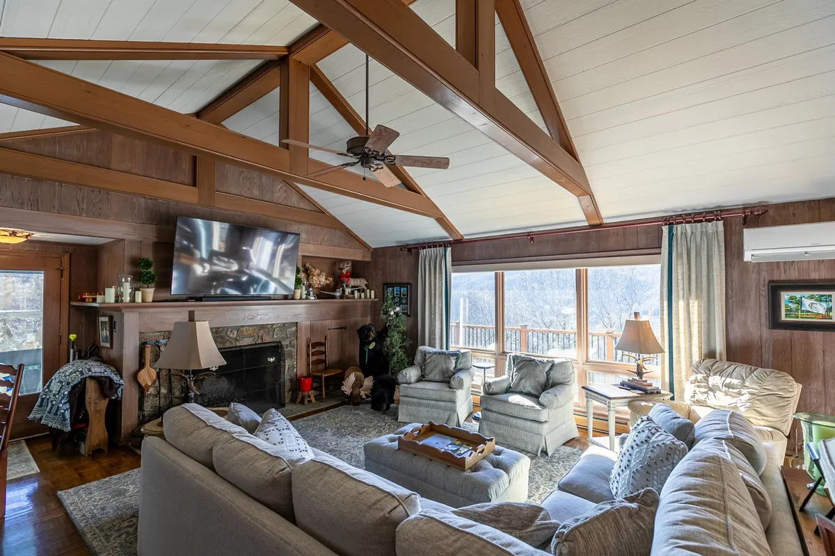 A spacious living room featuring a large sectional sofa positioned around a striking wood-burning fireplace. Expansive windows offer sweeping views of the surrounding mountains, while exposed wooden beams and a warm color palette create a welcoming ambiance. The setup invites both relaxation and social gatherings, with ample space for family and friends.