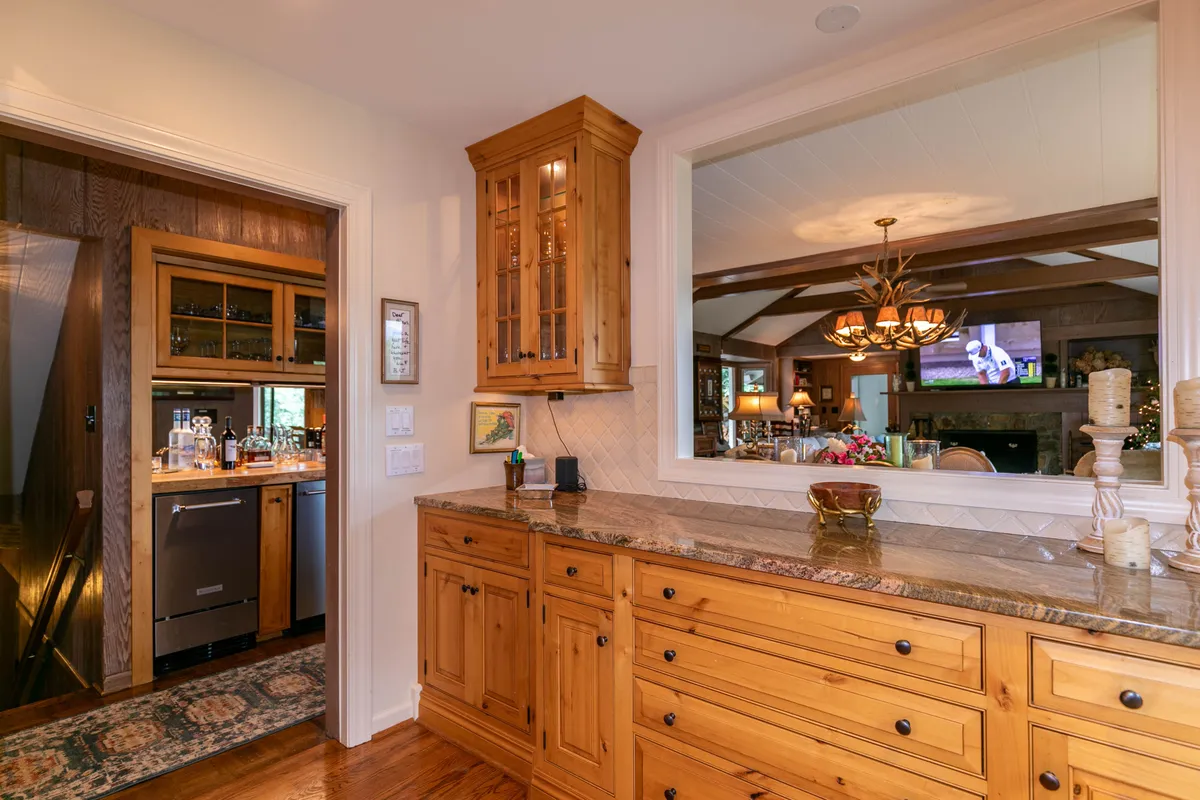 A warmly lit kitchen with classic cabinetry, expansive granite countertops, and a glass cabinet displaying dishware. The view into the adjacent living area hints at the open-plan layout, reinforcing the idea of the kitchen being connected to social spaces for group interaction.