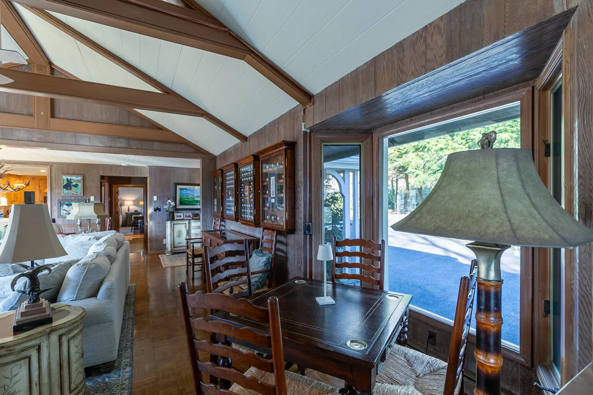 A dining area with a large wooden table set for meals, surrounded by comfortable seating. The room is bathed in natural light from floor-to-ceiling windows that reveal stunning mountain scenery outside. Wood accents and beams add rustic charm, while the inviting layout encourages togetherness and shared meals.