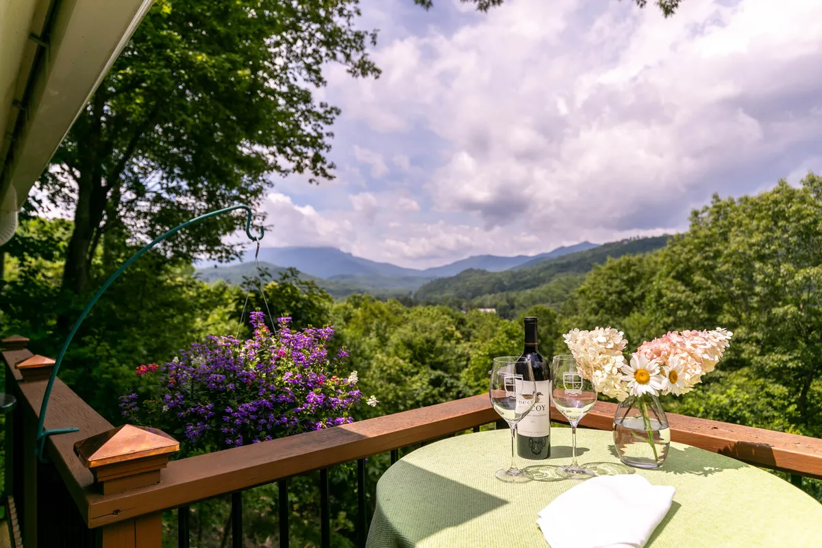 An inviting outdoor table set with wine glasses and flowers overlooks a stunning mountain view, surrounded by flowering plants. The setup hints at the perfect place for sunset toasts or relaxed group gatherings in the open air.