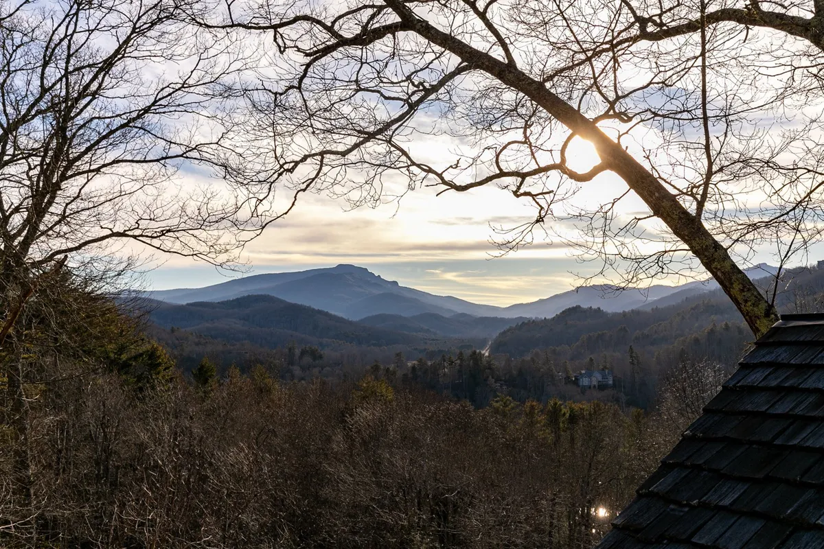 A sweeping view of a mountain range framed by silhouetted tree branches, with a cloudy sky and a distant valley below. The setting evokes the drama and beauty of North Carolina’s High Country, suggestive of hiking adventures and scenic vistas.