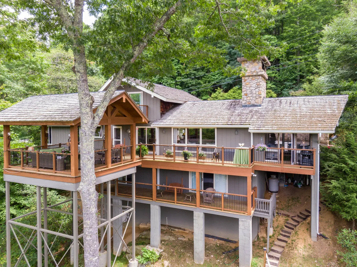 The large wooden deck wraps around the house, featuring multiple levels, a striking stone chimney, and is surrounded by mature trees. It promises space for gatherings, relaxation, and a direct connection to the natural environment.
