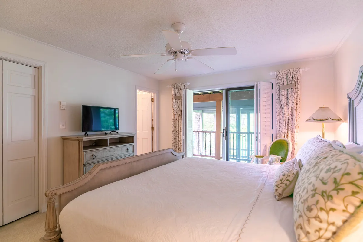 A serene king bedroom with decorative pillows, a television, an armchair, and a sliding glass door leading to a private balcony. Natural light fills the room, and the setup promises both relaxation and entertainment.