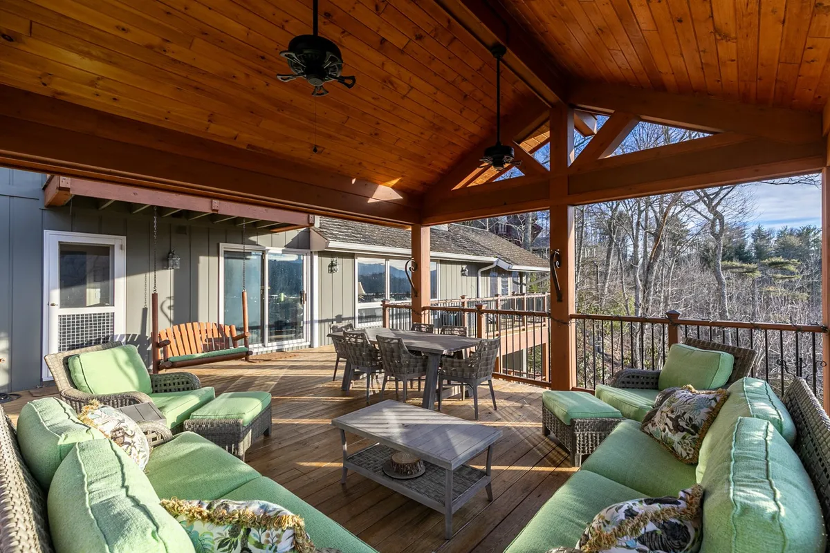 A spacious outdoor area with a green sectional sofa and dining table sits under a wooden ceiling, encircled by wooded greenery. The setup radiates a harmonious and relaxed vibe, ideal for socializing and enjoying meals together.